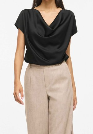 Femme portant un haut sans manches drapé en satin noir et un pantalon beige taille haute à jambes larges, debout devant un fond uni.