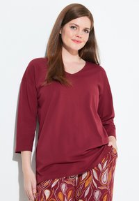 Vrouw met lang bruin haar, gekleed in een losse bordeauxrode top en gepatternde broek, staand tegen een witte achtergrond.