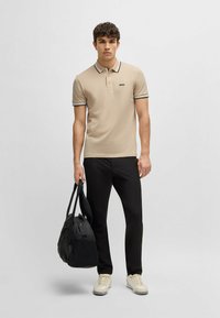 Beige polo skjorte med svart og hvit stripete krage, svarte bukser og beige joggesko. Bærer en svart stoffveske.