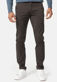 Mörkbruna chinos i mjukt tyg, slim-fit design, framfickor och kombinerade med vita sneakers. Subtil lyster och rena linjer.