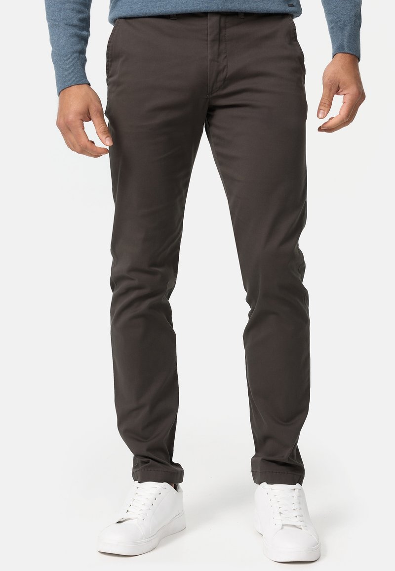 Mörkbruna chinos i mjukt tyg, slim-fit design, framfickor och kombinerade med vita sneakers. Subtil lyster och rena linjer.