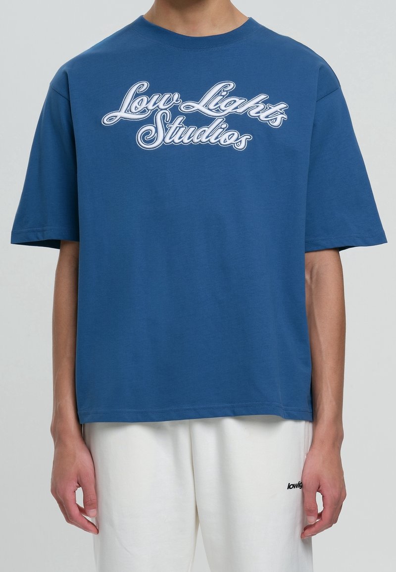 Personne portant un t-shirt bleu avec le texte "Low Lights Studios" et un pantalon blanc avec un petit texte noir sur la cuisse gauche.