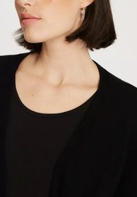 Czarny kardigan o miękkiej fakturze, z dekoltem w serek i otwartym przodem. Widać srebrne kolczyki oraz pasującą czarną bluzkę.