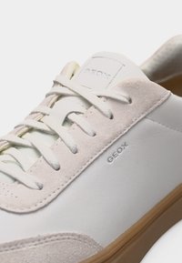 Λευκό αθλητικό παπούτσι Geox με σουέντ λεπτομέρειες, κορδόνια και φωτεινή καφέ λαστιχένια σόλα, εμφανίζεται σε κοντινή λήψη.