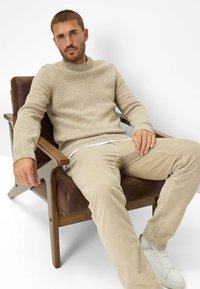 Beige gebreide trui met een tekstuurafwerking, gecombineerd met licht beige corduroy broek en witte sneakers, zittend in een houten fauteuil.