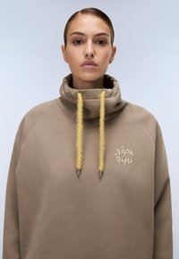 Maglione marrone con un collo alto a camino e cordini gialli in peluche. Presenta un logo sul petto. Tessuto liscio e strutturato.