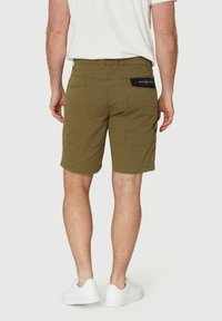 Bermudas marrones de corte relajado, con un bolsillo trasero que tiene un parche negro con el logotipo y detalles de costura. Usadas con zapatillas blancas.