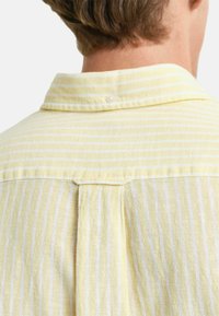 Camicia con righe gialle e bianche, presenta un colletto button-down e un dettaglio di piega sul retro. Il tessuto appare morbido e testurizzato.