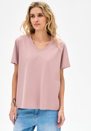 Blonde vrouw met golvend haar draagt een licht mauve V-hals top met korte mouwen en lichtblauwe spijkerbroek tegen een effen witte achtergrond.