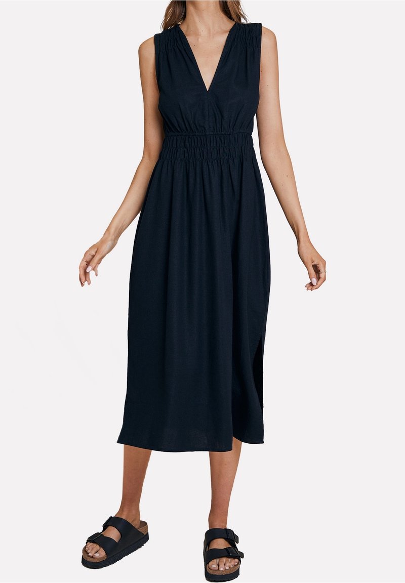 Marineblaues ärmelloses Kleid mit V-Ausschnitt, elastischem Bund und seitlichen Schlitzen. Der Stoff ist leicht und hat eine glatte Textur. Kombiniert mit schwarzen Sandalen.
