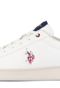Baskets blanches avec semelle crème, lacets blancs, languette bleu marine au talon et logo rouge représentant deux joueurs de polo à cheval sur le côté.