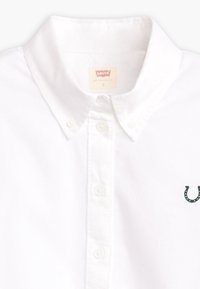 Chemise blanche en coton boutonnée avec un col à revers, ornée d'un petit logo vert brodé sur la poitrine gauche.