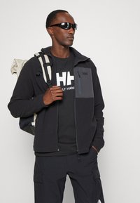 Veste en polaire noire avec panneau gris, poche zippée, associée à un t-shirt noir avec un grand logo blanc, un pantalon cargo et un sac à dos.