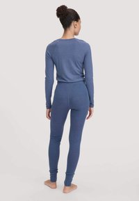 Vrouw die blootvoets met de rug naar ons toe staat, gekleed in een strakke blauwe lange mouwen top en bijpassende leggings, tegen een effen achtergrond.