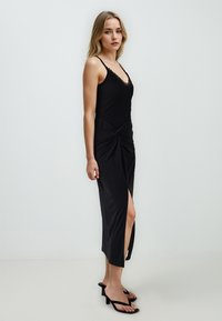 Zwarte jurk met spaghettibandjes, met een geknoopte design bij de taille en een zijsplit. Gladde, lichte stof. Hakken sandalen.