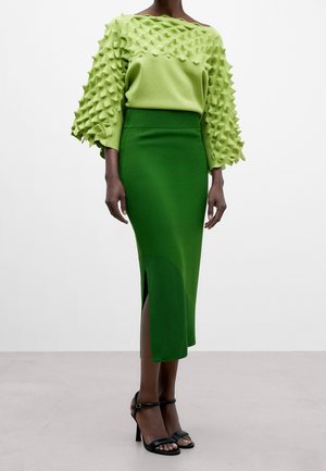 Person iført en limegrøn tekstureret langærmet top og en mørkegrøn højtaljet midinederdel med en frontslids, kombineret med sorte sandaler med hæl.