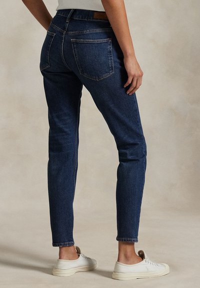 Osoba oblečená v tmavomodrých úzkych rifliach a bielych teniskách stojaca s jednou rukou voľne pozdĺž tela.