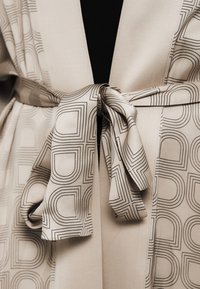 Robe en tissu beige avec motif géométrique noir, dotée d'une coupe portefeuille et d'une taille nouée. Texture lisse avec une apparence fluide.