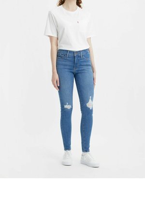 Kobieta w białym t-shircie z krótkim rękawem, niebieskich dopasowanych jeansach z wysokim stanem i przetarciami na kolanach oraz białych trampkach, stojąca na jednolitym tle.