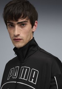 Sort lynlåsjakke med høj krave, præget af hvid "PUMA"-logo og detaljer. Fremstillet af glat, strækbart stof med minimal tekstur.
