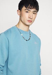 Světlomodrá mikina s kulatým výstřihem z bavlněné směsi. Obsahuje malé bílé logo Nike a kovový řetízkový náhrdelník jako doplněk.