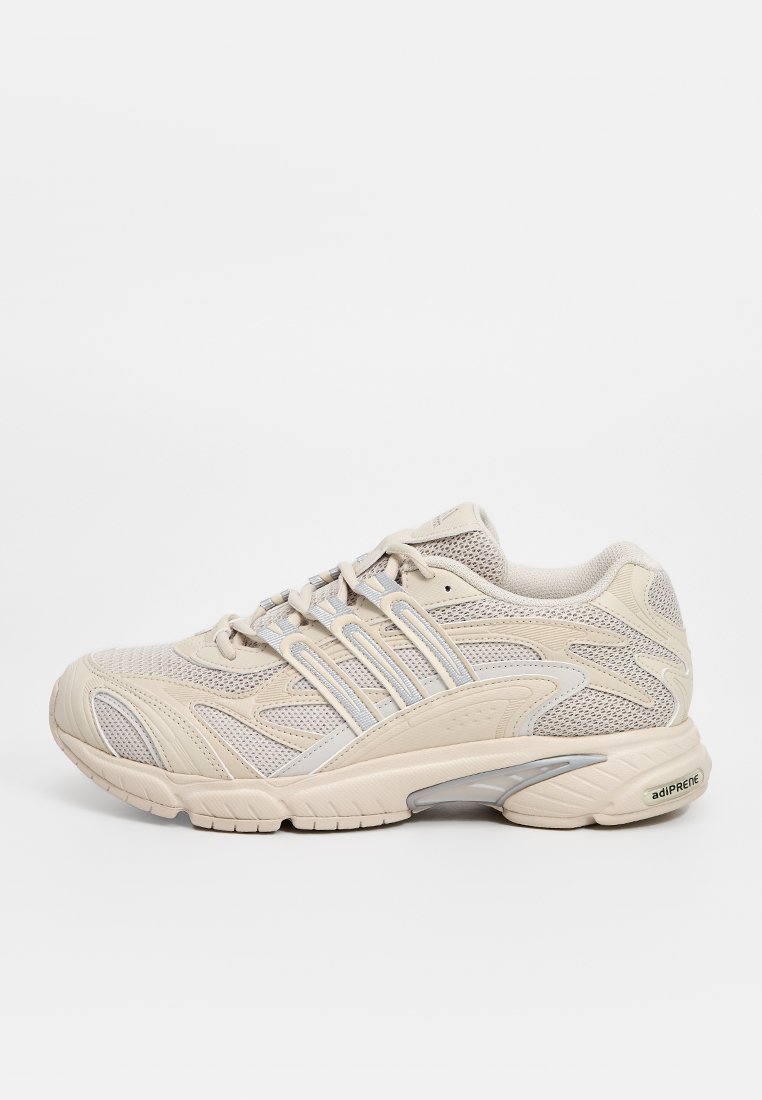 Zapatilla deportiva beige con parte superior de malla, acentos plateados y suela texturizada. Cuenta con un sistema de cordones y un acolchado prominente para mayor soporte.