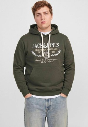 Un homme porte un sweat à capuche Jack & Jones vert foncé, avec des cordons blancs et une inscription sur la poitrine, associé à un jean bleu clair.