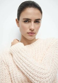 Pull tricoté rose clair avec un design ample et texturé et de larges poignets côtelés, porté par un mannequin sur un fond neutre.
