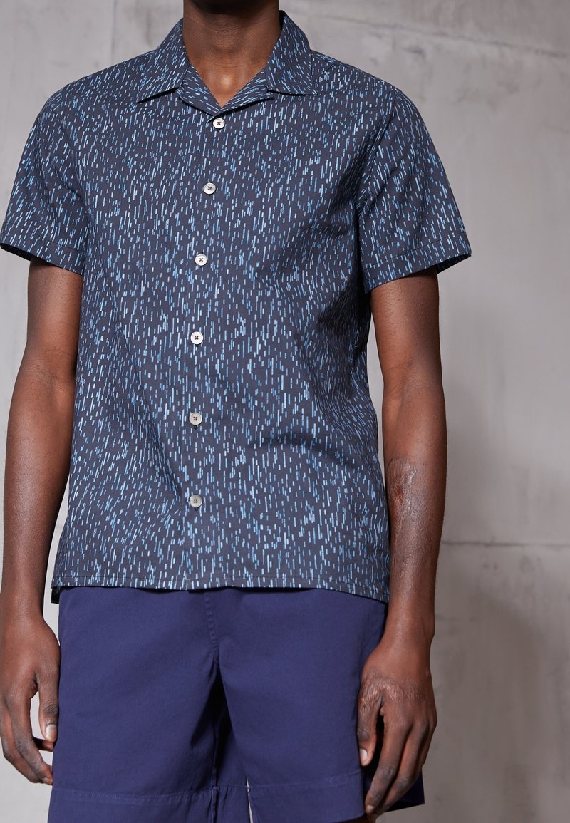 Mężczyzna w granatowej koszuli z krótkim rękawem, zapinanej na guziki, z jasnoniebieskim wzorem oraz ciemnoniebieskich szortach na jednolitym tle.