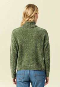 Grön, texturerad stickad tröja med hög krage och sänkt axlar. Ribbad mudd och nederkant, ihop med blå högmidjade jeans.