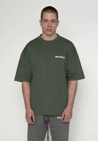 Grøn oversized t-shirt i bomuld med korte ærmer, med en hvid "MULTIPLY"-logo på venstre brystområde, stylet med grå sweatpants.