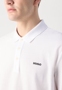 Hvit polo skjorte laget av bomull, med klassisk krage, tre-knaps placket, og en svart "HUGO" logo på venstre side.