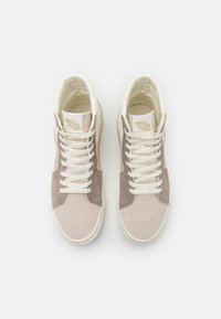 Zapatillas de caña alta en ante de color beige claro y topo, con cordones blancos, un cuello acolchado y detalles de costura sutiles en la puntera.
