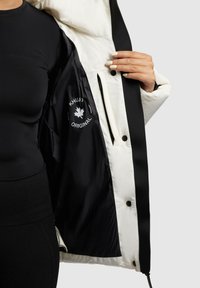 Vooderdatud jope omab kreemjat väliskihti musta voodriga, mille sees on logopatch. Nähtavad nuppude ja nööriga reguleerimise võimalused.