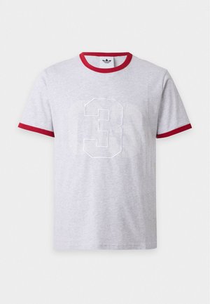 Jeune homme debout portant un T-shirt gris avec un liseré rouge et un grand numéro trois blanc sur le devant, associé à un pantalon noir.