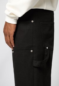 Pantalon en denim noir avec une poche latérale, des accents métalliques argentés et une finition texturée, posé à côté d'une manche blanche texturée.