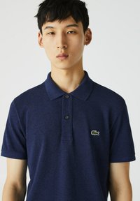 Polo en marine avec un tissu texturé, deux boutons noirs et un logo de crocodile brodé en vert sur la poitrine. Manches courtes.