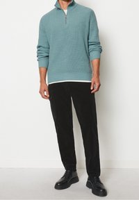 Pull côtelé bleu sarcelle avec col à demi-fermeture éclair, porté sur une chemise blanche, accompagné d'un pantalon en velours côtelé noir et de chaussures noires à lacets.