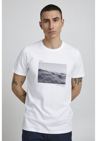 Hvid bomulds-t-shirt med et grafisk print af bjerge, med vandrette linjer og tekst nedenfor. Rund halsudskæring, korte ærmer.