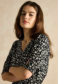 Schwarzes Kleid mit weißem Blumenmuster, V-Ausschnitt, kurzen Volantärmeln, gerafften Bündchen und strukturiertem Stoff. Modell mit verschränkten Armen.