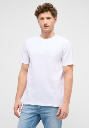 Junger Mann mit lockigem Haar, der ein weißes Henley-Shirt mit kurzen Ärmeln und hellblaue Jeans trägt, steht vor einem einfarbigen Hintergrund.