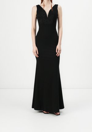 Woman wearing a sleeveless black evening gown with lace bodice and deep V neckline, standing with hands relaxed by sides.
