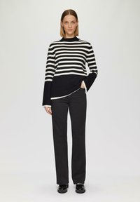 Gestreifter Pullover in Schwarz-Weiß mit einem Stehkragen, der bis zur Hüfte reicht, kombiniert mit schwarzen Hosen und glänzenden schwarzen Schuhen.