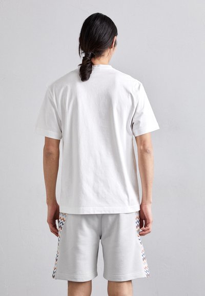 T-shirt en coton blanc à manches courtes, associé à un short gris clair avec des motifs en zigzag bleus, oranges et jaunes sur les côtés.