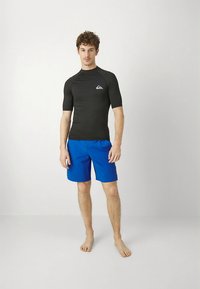 Svart kortärmad rashguard med logotyp, kombinerad med blå badshorts. Materialet verkar vara stretchigt och mjukt, lämpligt för vattenaktiviteter.