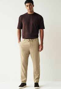 T-shirt marron à manches courtes associé à un pantalon beige avec une taille élastique à cordon, et des baskets noires sur un fond uni.