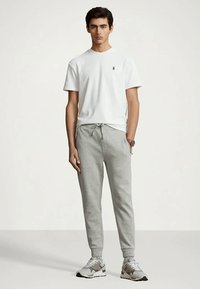 Man som står klädd i en vit t-shirt med korta ärmar, ljusgrå joggingsbyxor och grå sneakers framför en enfärgad bakgrund.