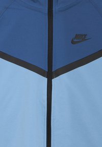 Μπουφάν Nike με χρωματικό συνδυασμό μπλε και ανοιχτό μπλε, μαύρο φερμουάρ και λογότυπο, υφασμάτινη υφή και σχέδια με σχήματα σφήνας.