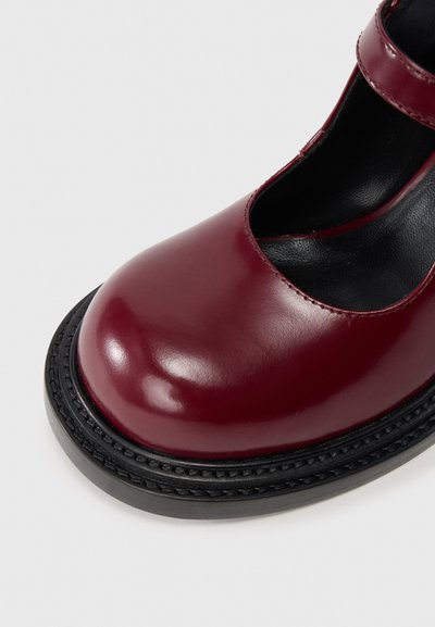 Bordowe lakierowane buty typu Mary Jane z okrągłym noskiem, czarną teksturowaną podeszwą i cienkim czarnym paskiem na wierzchu stopy.
