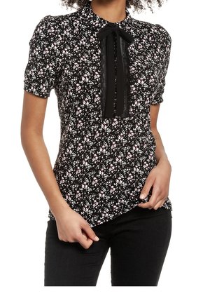 Kobieta nosząca czarną krótki rękaw kwiatową bluzkę z małymi białymi i różowymi kwiatami, zapinana z przodu na guziki, z czarną kokardą przy kołnierzyku.